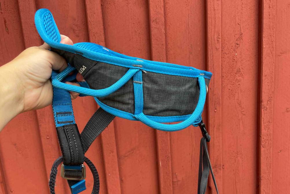 Klettergurt-Erwachsene-Test: Ein blau-schwarzer Klettergurt von Salewa wird vor einer roten Holzwand in der Hand gehalten. 