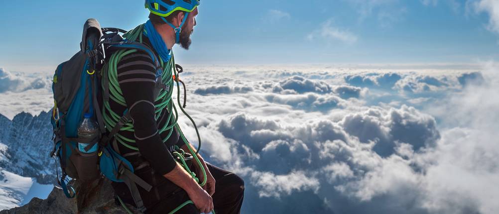 Kletterhelm im Test: Mann mit blauem Kletterhelm sitzt auf einem Felsbrocken an der Spitze des Berges und schaut auf die Wolken herab.
