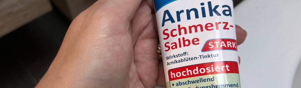 Arnika-Salben im Test: Hand hält eine Tube mit Arnika-Schmerzsalbe von Klosterfrau vor einer weißen Fläche.