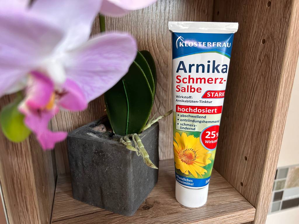 Arnika-Salbe von Klosterfrau im Test: eine Tube steht in einem Holzregal neben Blumen.