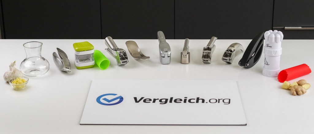 Verschiedene Knoblauchpressen liegen in einem Halbkreis auf einer weißen Tischplatte, vor einem Schild mit dem Schriftzug Vergleich.org.