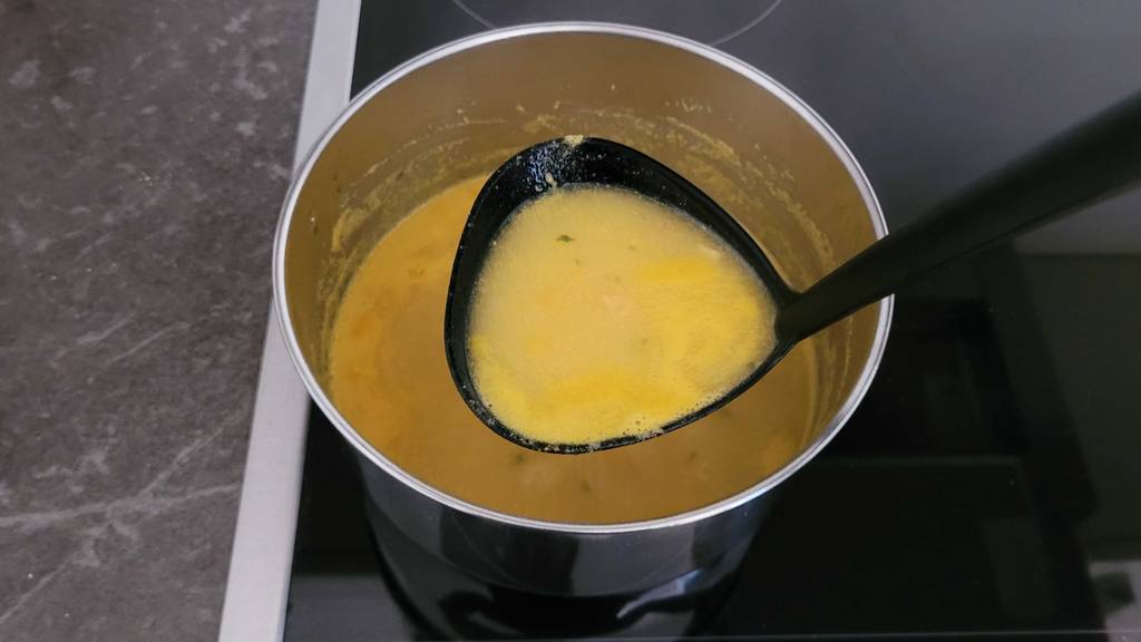 Eine Suppenkelle mit getesteter Kürbissuppe über einem von oben betrachteten Topf, der auf einer Herdplatte steht.