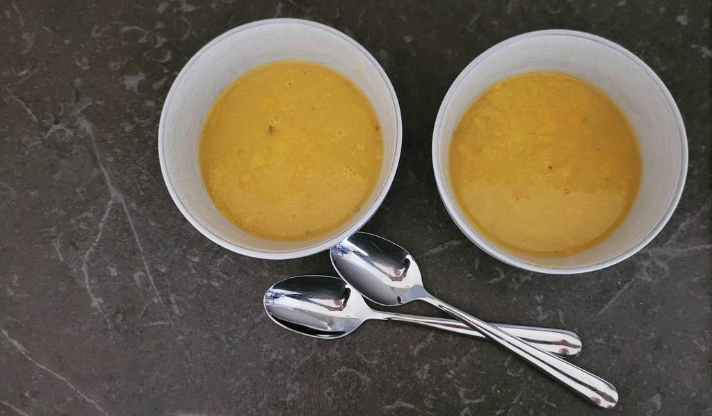 Ansicht von oben auf zwei mit getesteter Kürbissuppe gefüllte weiße Suppenschalen, die neben zwei Esslöffeln auf einer schwarzen Oberfläche stehen.