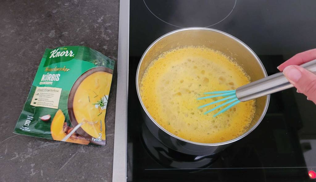 Eine Hand hält einen Schneebesen in einen von oben betrachteten Topf inklusive kochender, getesteter Kürbissuppe, der auf einer Herdplatte neben einer Verpackung Knorr Feinschmecker Kürbis-Cremesuppe steht.