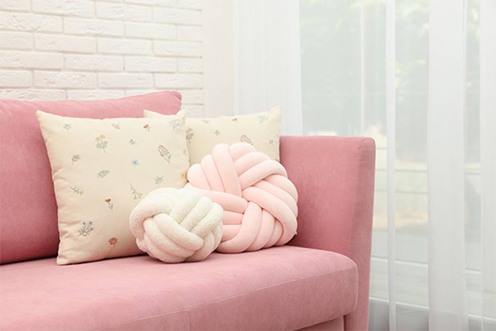 Knotenkissen im Test: Ein großes rosa Knotenkissen und ein kleineres weißes auf einer rosa Couch mit weiteren Kissen.