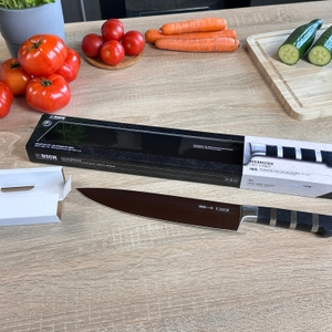 Das F.-Dick-1905 Kochmesser liegt neben geöffneter Verpackung und Kartoneinlage auf dem Tisch. Gemüse ist im Hintergrund sichtbar.