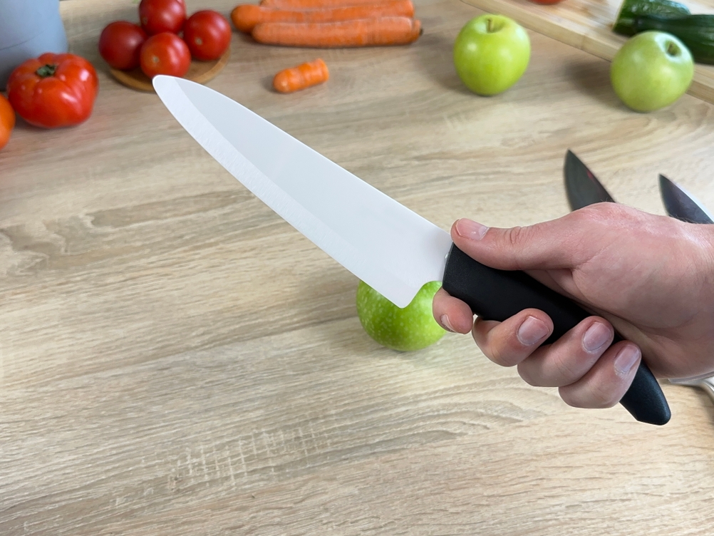 Das Kyocera-Keramikmesser wird in der Hand gehalten. Im Hintergrund sind Gemüse und ein Apfel sichtbar.