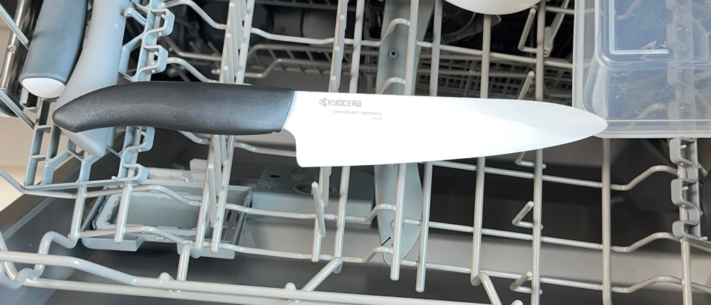 Ein Kyocera-Keramikmesser liegt im oberen Korb einer geöffneten Spülmaschine. Weitere Küchenutensilien sind ebenfalls einsortiert.