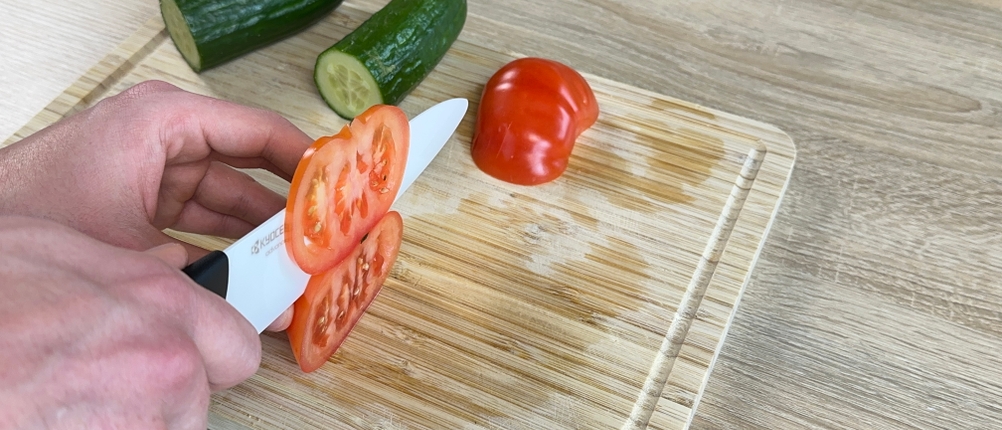 Das Kyocera-Keramikmesser schneidet eine Tomate auf einem Holzbrett. Die Hand führt den Schnitt von oben.