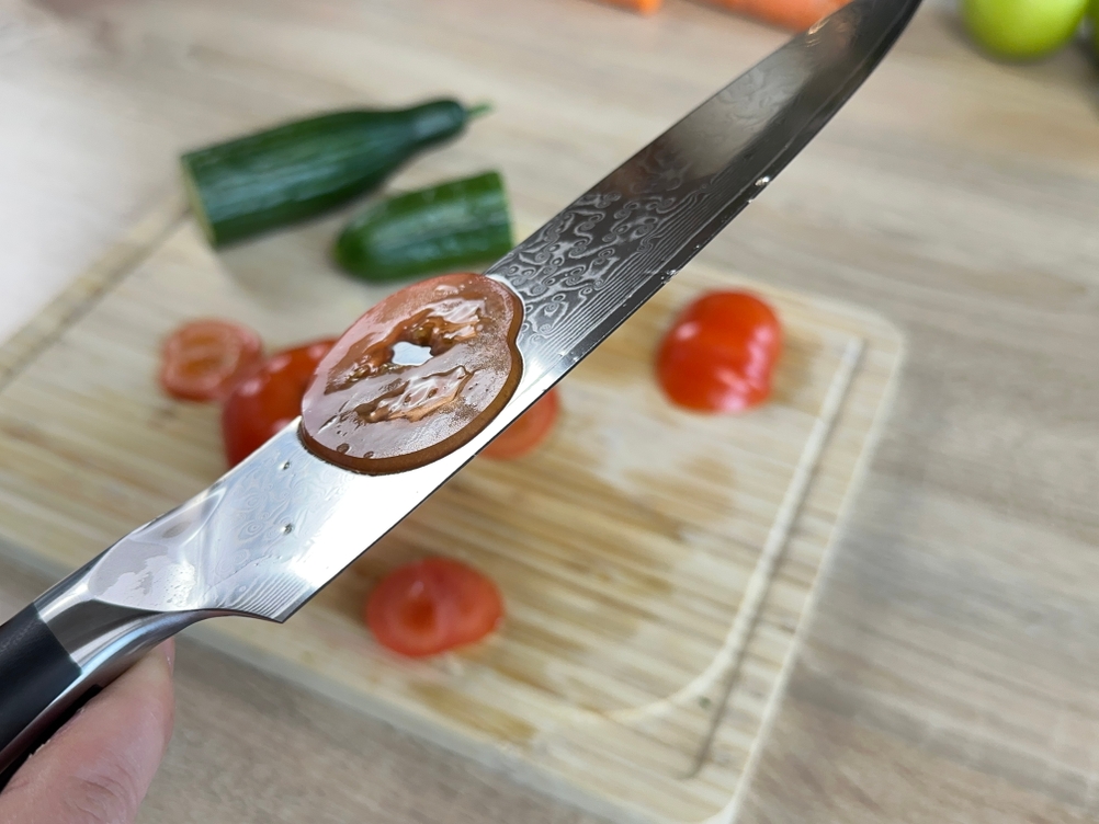 Eine extrem dünne Tomatenscheibe haftet an der Klinge des Shan-Zu-Messers. Das Schneidebrett liegt im Hintergrund.