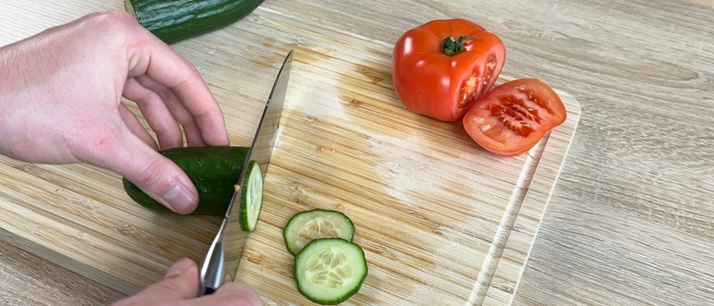 Das Shan-Zu-Kochmesser wird beim Schneiden einer Gurke auf einem Holzbrett benutzt. Eine Tomate liegt daneben.