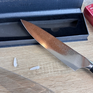 Nahaufnahme des Shan-Zu-Kochmessers mit detaillierter Damaststruktur. Die geöffnete Verpackung liegt daneben.