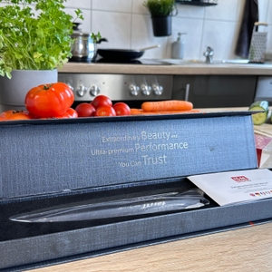 Das Shan-Zu-Kochmesser liegt geöffnet in seiner schwarzen Verpackung mit Benutzerhandbuch und Klingenschutz. Im Hintergrund stehen Tomaten.