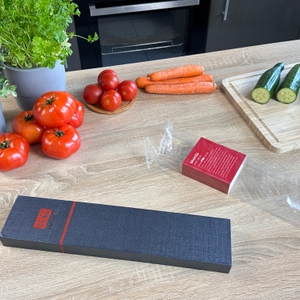 Die geschlossene Verpackung des Shan-Zu-Kochmessers liegt auf einer Arbeitsfläche. Gemüse und Zubehör liegen im Hintergrund.