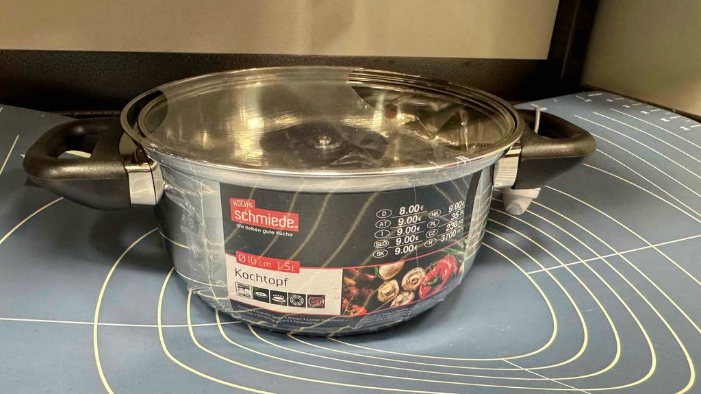 Ein in Plastik eingeschweißter, getesteter Kochtopf 16 cm steht auf einer Backfolie aus grauem Silikon.