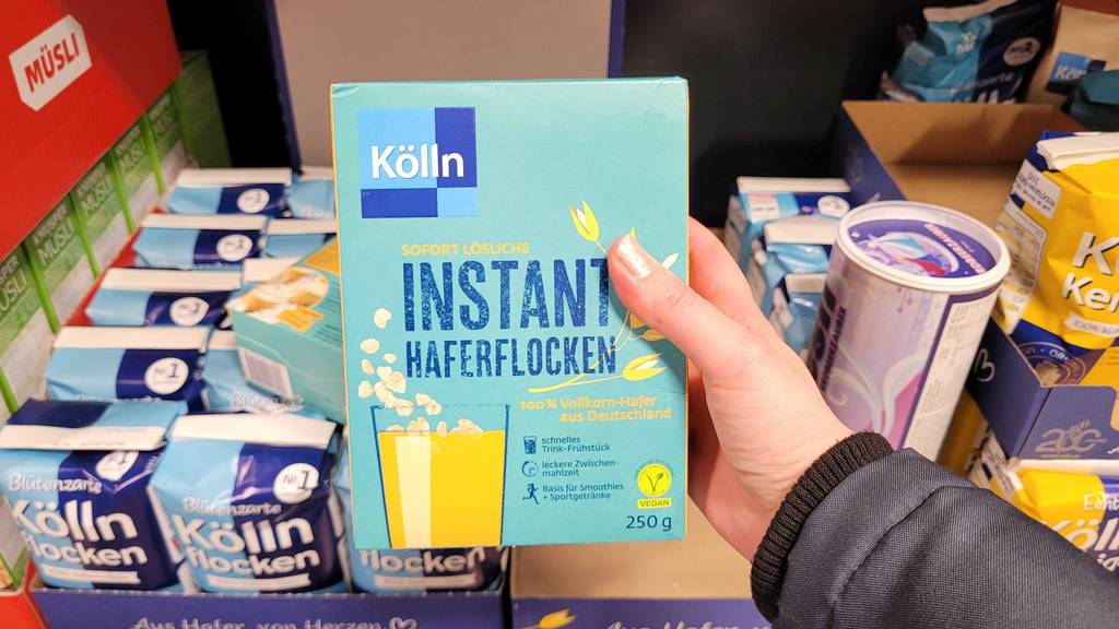 Eine Damenhand hält eine 250-g-Verpackung mit Instant Haferflocken im Test vor einem Lebensmittelregal.