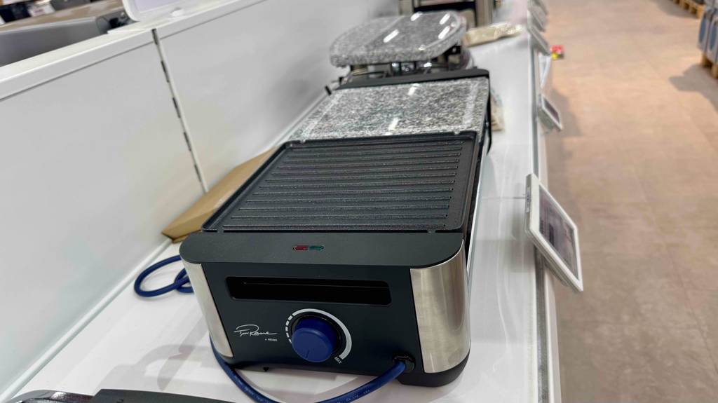 Der elektrische getestete Raclette-Grill mit Steinplatte steht in einer Reihe mit weiteren Grillmodellen auf einem Ausstellungsregal im Geschäft.
