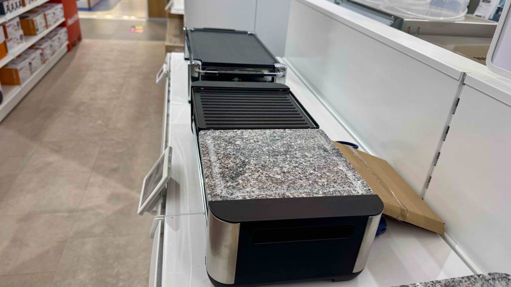 Seitenansicht auf einen Raclette-Grill mit Steinplatte im Test, der inklusive Marmor-Platte auf einem Ausstellungsregal neben weiteren Grill-Geräten steht.