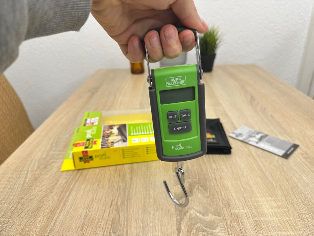 Eine Burg-Wächter-Tara-PS-7600-Kofferwaage wird am Griff gehalten, während der Haken zur Befestigung eines Koffers bereitsteht.