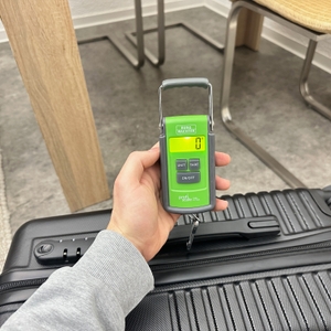 Eine Burg-Wächter-Tara-PS-7600-Kofferwaage misst das Gewicht eines schwarzen Hartschalenkoffers und zeigt den Wert auf dem grün beleuchteten Display an.