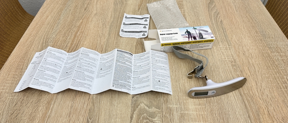 Der Lieferumfang einer Mycarbon-DS2-Kofferwaage liegt vollständig ausgepackt auf einem Holztisch mit Waage, Trageband, Anleitung und Verpackung.