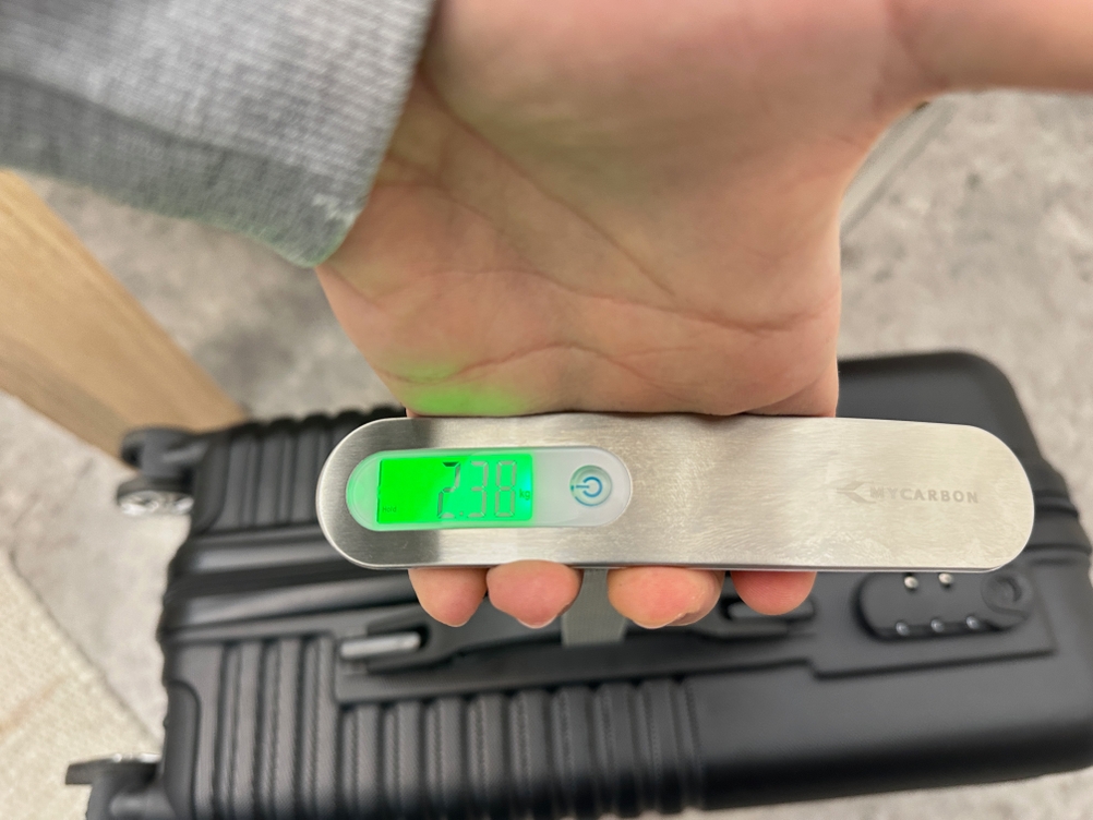 Eine Mycarbon-DS2-Kofferwaage wird über ein Trageband an einem Koffer befestigt, während das Display das Gewicht anzeigt.