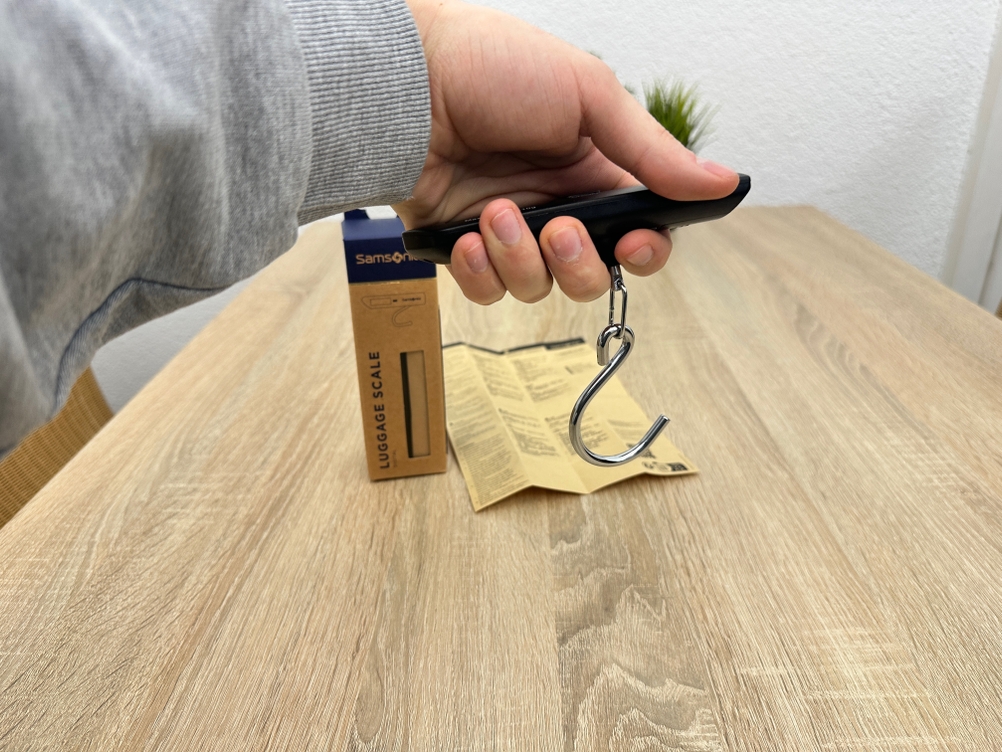 Eine Samsonite-122291/1041-Kofferwaage wird von einer Hand gehalten, während der Metallhaken frei nach unten hängt.