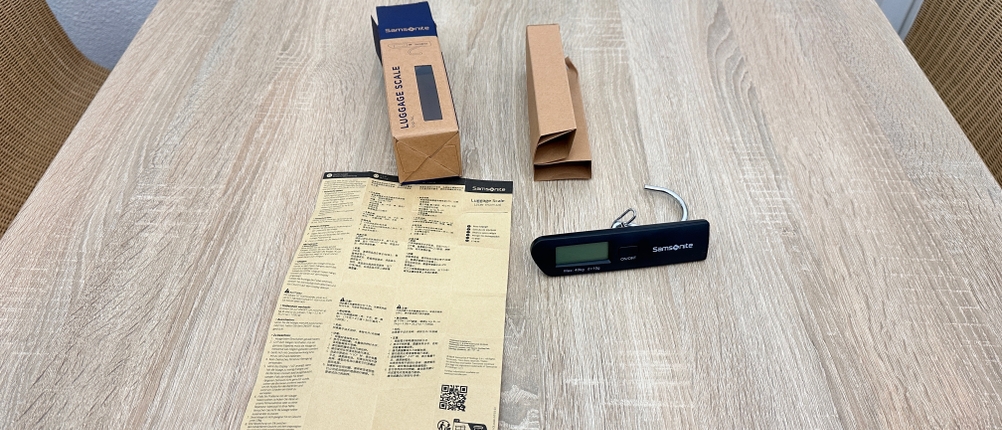 Der Lieferumfang einer Samsonite-122291/1041-Kofferwaage liegt mit Verpackung, Anleitung und der schwarzen Digitalwaage auf dem Tisch.