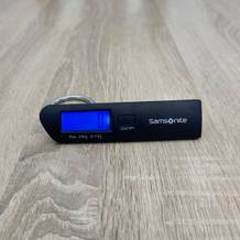 Eine Samsonite-122291/1041-Kofferwaage liegt eingeschaltet auf einem Holztisch, während das blaue Display aktiv ist.