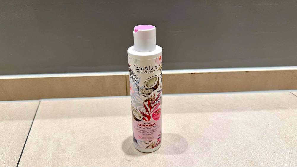 Auf einem hellen Boden vor einer grauen Wand steht eine Flasche des getesteten Kokos-Shampoos.