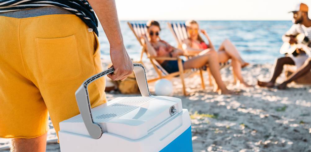 Ein Mann hält eine Kühlbox, im Hintergrund warten Freunde am Strand