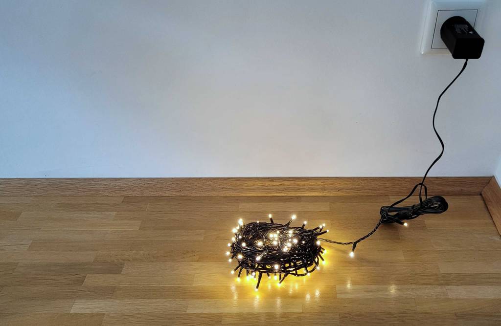 Eine LED-Weihnachtsbeleuchtung liegt aufgerollt und in der Steckdose zum testen angesteckt auf einem Laminatboden.
