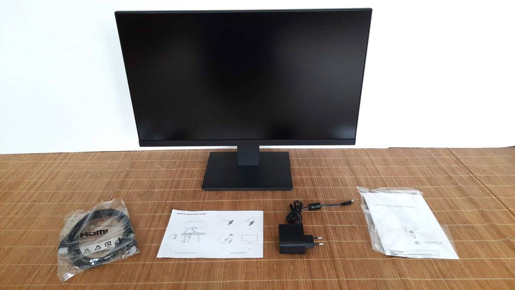 Vorderansicht eines schwarzen Koorui 24-Zoll-Monitors im Test, der inklusive Zubehör, wie Kabel und Gebrauchsanleitung, auf einer hellbraunen Bastmatte steht.