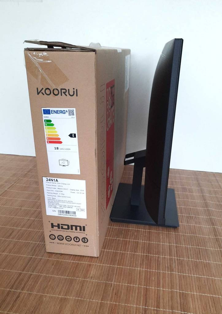 Seitenansicht eines Koorui 24-Zoll-Monitors im Test, der neben seinem braunen Originalverpackungskarton auf einer hellbraunen Bastmatte steht.