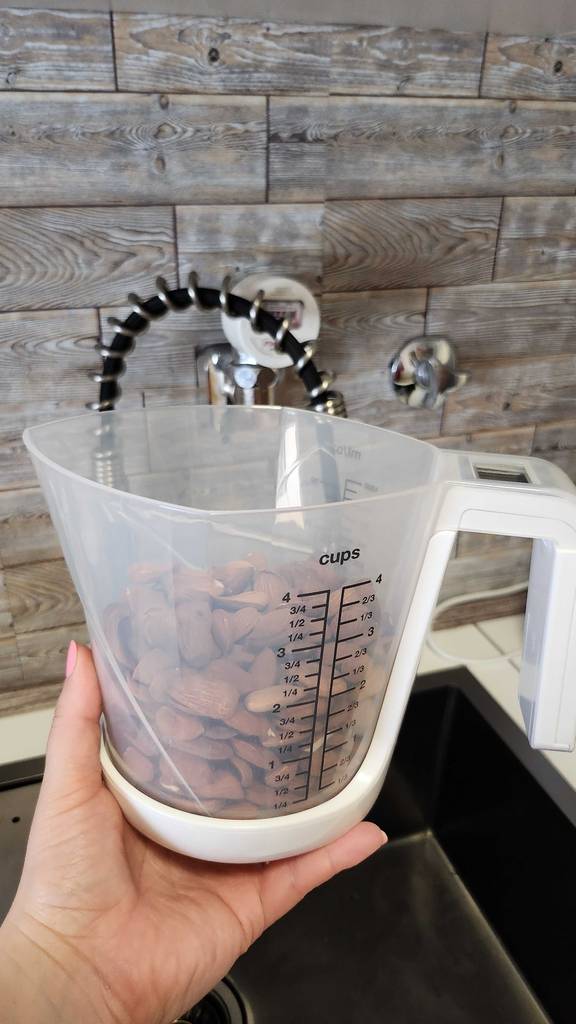 Eine Hand hält einen getesteten Messbecher mit Waage, in dem sich Mandeln befinden, über eine Küchenspüle.