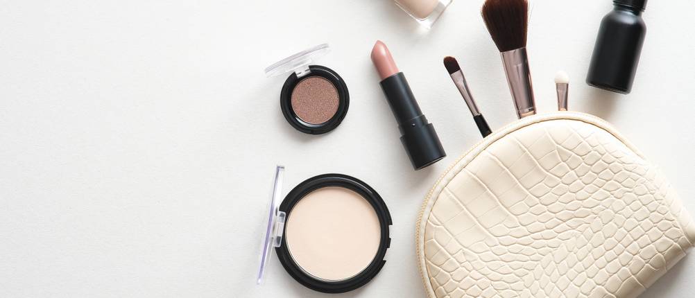Kosmetiktasche-Test: Auf weißem Hintergrund liegen Make-up, Pinsel und Lippenstift. Daneben liegt eine helle kleine Tasche.