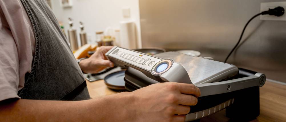 Mann mit Schürze bedient einen Elektrogrill auf der Küchenarbeitsfläche.