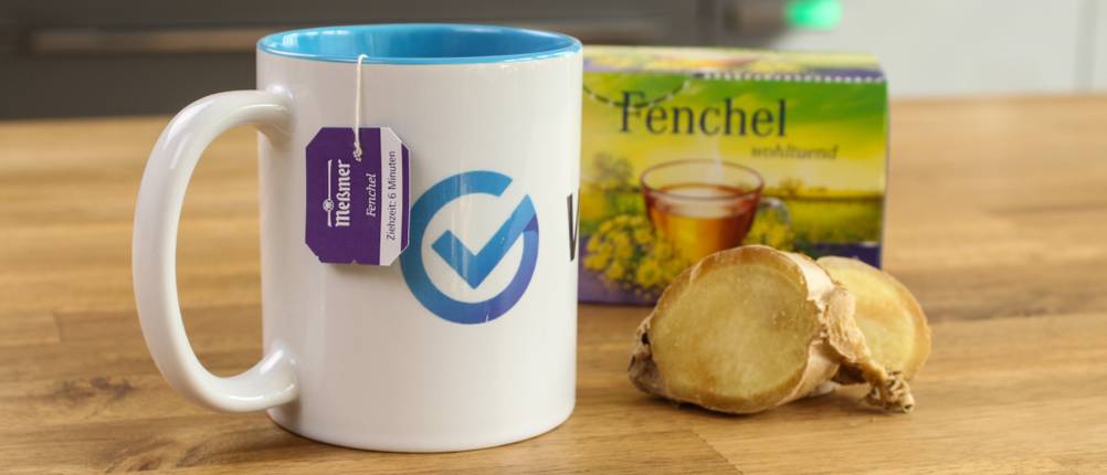 Kräutertee-Test: Auf einem Tisch steht ein Glas mit Tee. Daneben befinden sich eine Ingwerknolle und eine Tee-Verpackung.