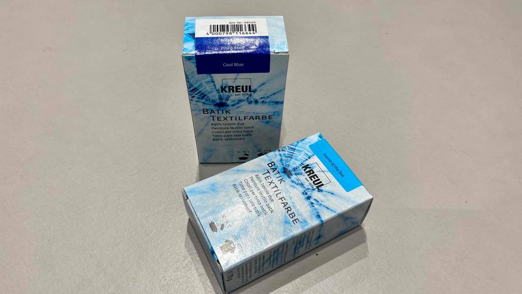 Eine getestete Batikfarbe in Cool Blue steht in der Verpackung neben einer liegenden Schachtel mit Sound of the Sea Farbton auf einer hellen Oberfläche.