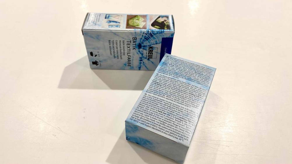 Batikfarben-Test: zwei Verpackungen mit Sicht auf die Vorderseite sowie die Rückseite inklusive Anwendungsanleitung liegen beieinander auf einem hellen Untergrund.