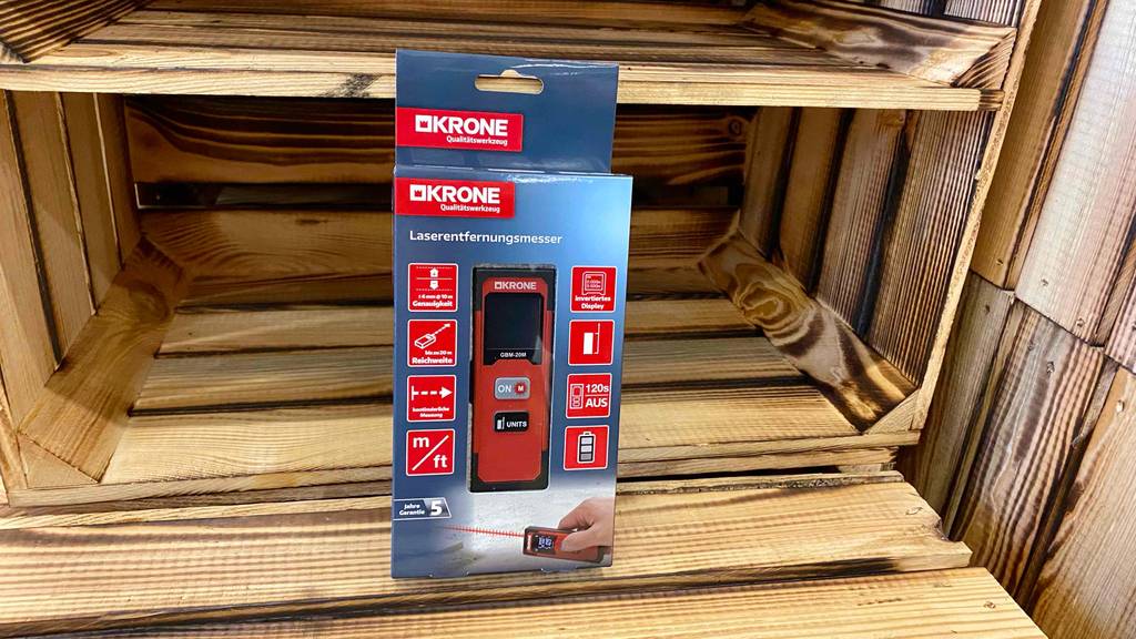 Ein verpacktes Laser-Entfernungsmesser der Marke Krone steht zum testen vor einer Holzkiste.