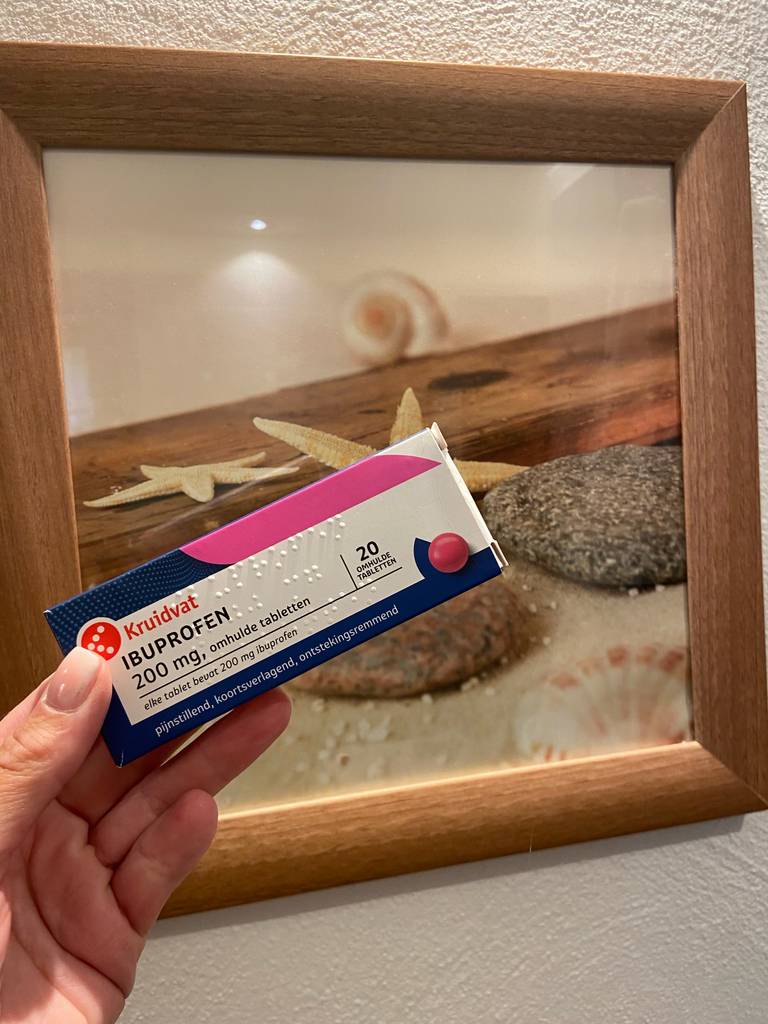 Eine Hand hält das getestete Kopfschmerzmittel in der Packung mit zwanzig Ibuprofen-Tabletten vor einem Bild mit Muschelmotiv.