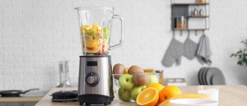 Krups-Standmixer im Test: Ein mit Obst gefüllter Standmixer steht in einer hellen Küche auf einer Holzarbeitsfläche. Daneben sind eine Reihe von Obst und eine weiße Schale sowie ein Holzbrett mit Messer zu sehen.