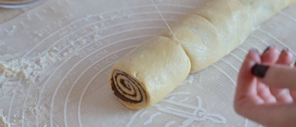 Nahansicht von Frauenhänden, die mit Küchengarn eine Rolle Zimtschneckenteig schneiden.