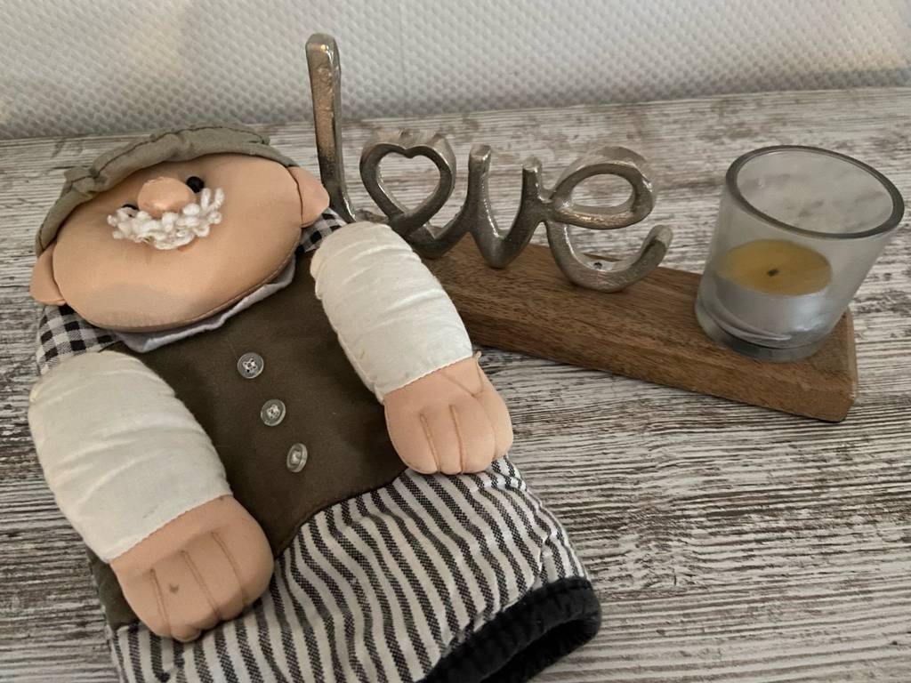 Getestet: Der Küchenhandschuh aus schwarz-weiß kariertem Stoff, mit darauf genähter, ausgestopfte Puppe, liegt auf einem Tisch mit Dekokerze.