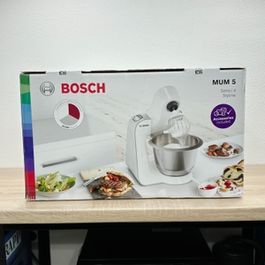 Die Verpackung zeigt die Bosch MUM Serie 4 Styline in Weiß. Rund um die Küchenmaschine sind servierte Speisen abgebildet.