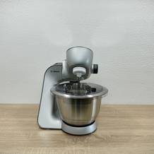 Die Bosch MUM56340 steht komplett aufgebaut auf einer Küchenfläche. Die Rührschüssel ist eingesetzt, der Spritzschutz montiert.