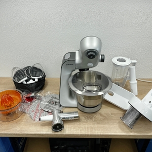 Die Bosch MUM56340 ist umgeben von Zubehörteilen wie Rührbesen, Entsafter, Reibe und Mixeraufsatz. Alles liegt auf einer Arbeitsfläche.