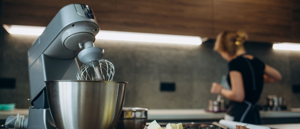 Küchenmaschinen-Test: Auf einer Küchenarbeitsfläche steht eine Küchenmaschine und daneben zwei Schalen mit weißer und Vollmilch-Schokolade. Im Hintergrund steht eine Frau.