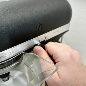 Ein Handgriff betätigt den Sicherungshebel der KitchenAid Artisan, um den Motorkopf zu entriegeln. Der Mechanismus ist gut sichtbar.